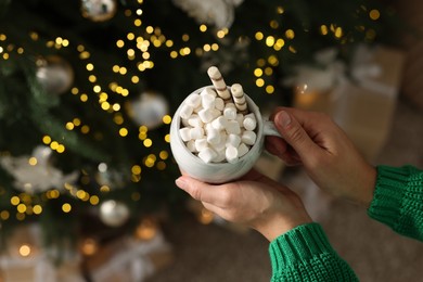 Woman holding cup of delicious Christmas cocoa with marshmallows and wafer sticks near fir tree, above view