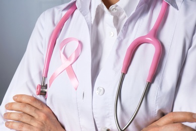 Female doctor with pink ribbon and stethoscope on grey background, closeup. Breast cancer concept