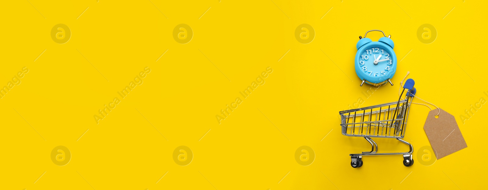 Photo of Shopping cart with tag, alarm clock  and space for text on yellow background, flat lay. Sale concept