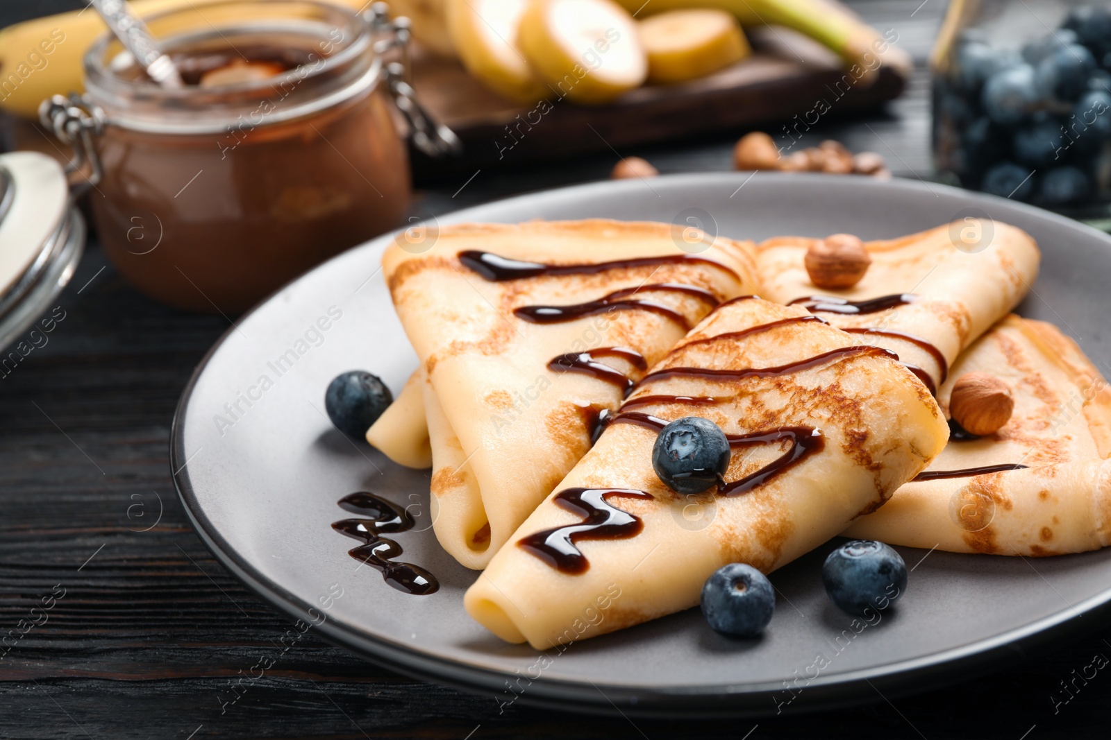 Photo of Delicious thin pancakes with chocolate, blueberries and nuts on black wooden table