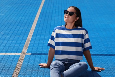 Beautiful woman in sunglasses on blue floor covering outdoors, space for text