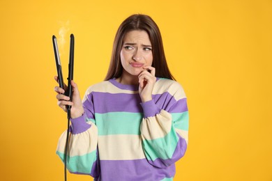 Photo of Upset young woman with flattening iron on yellow background. Hair damage