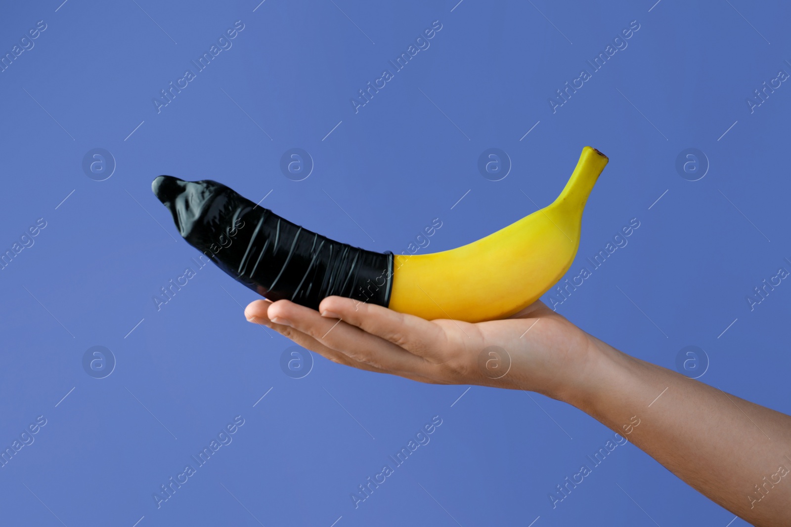 Photo of Woman holding banana in condom on light blue background, closeup. Safe sex concept