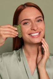 Young woman massaging her face with jade gua sha tool on green background