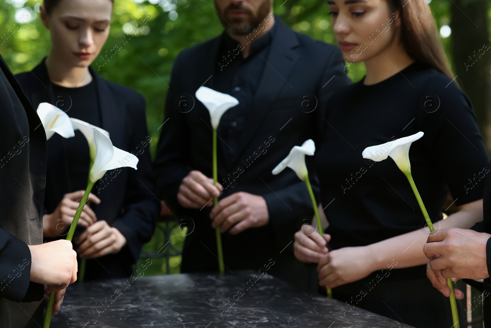 Photo of People with calla lily flowers near granite tombstone at cemetery outdoors, closeup. Funeral ceremony