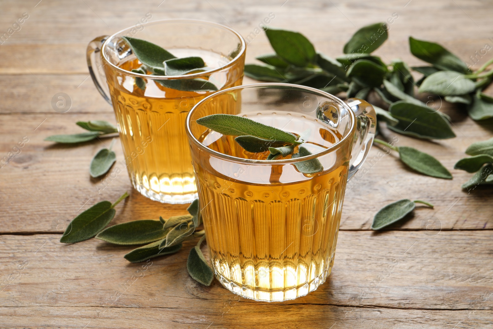 Photo of Cups of aromatic sage tea and fresh leaves on wooden table