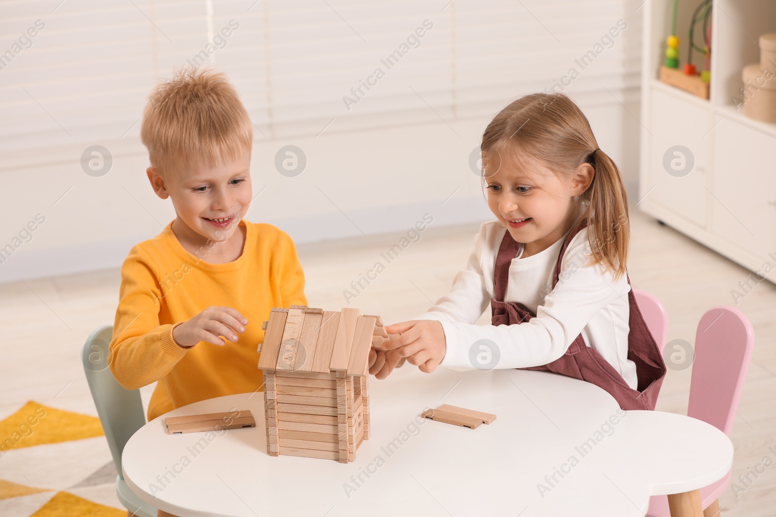 Photo of Little boy and girl playing with wooden house at white table indoors. Children's toys