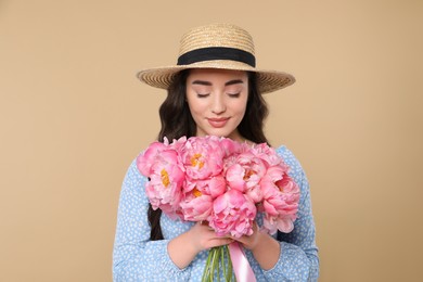 Photo of Beautiful young woman in straw hat with bouquet of pink peonies against beige background