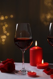 Glasses of red wine, burning candles and rose flower on grey table against blurred lights. Romantic atmosphere