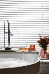 Photo of White wooden tray with glass of rose wine, book and burning candles on bathtub in bathroom