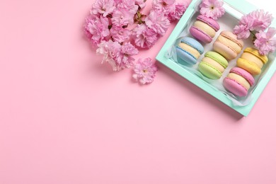 Photo of Many delicious colorful macarons in box and flowers on pink background, flat lay. Space for text