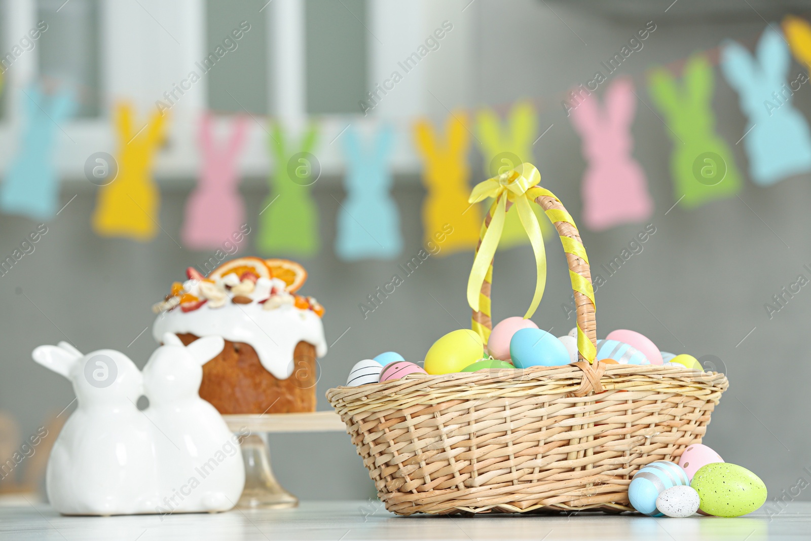 Photo of Easter basket with tasty painted eggs near cake and figure of rabbits on white table indoors