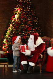 Photo of Santa Claus giving present to little boy near Christmas tree indoors