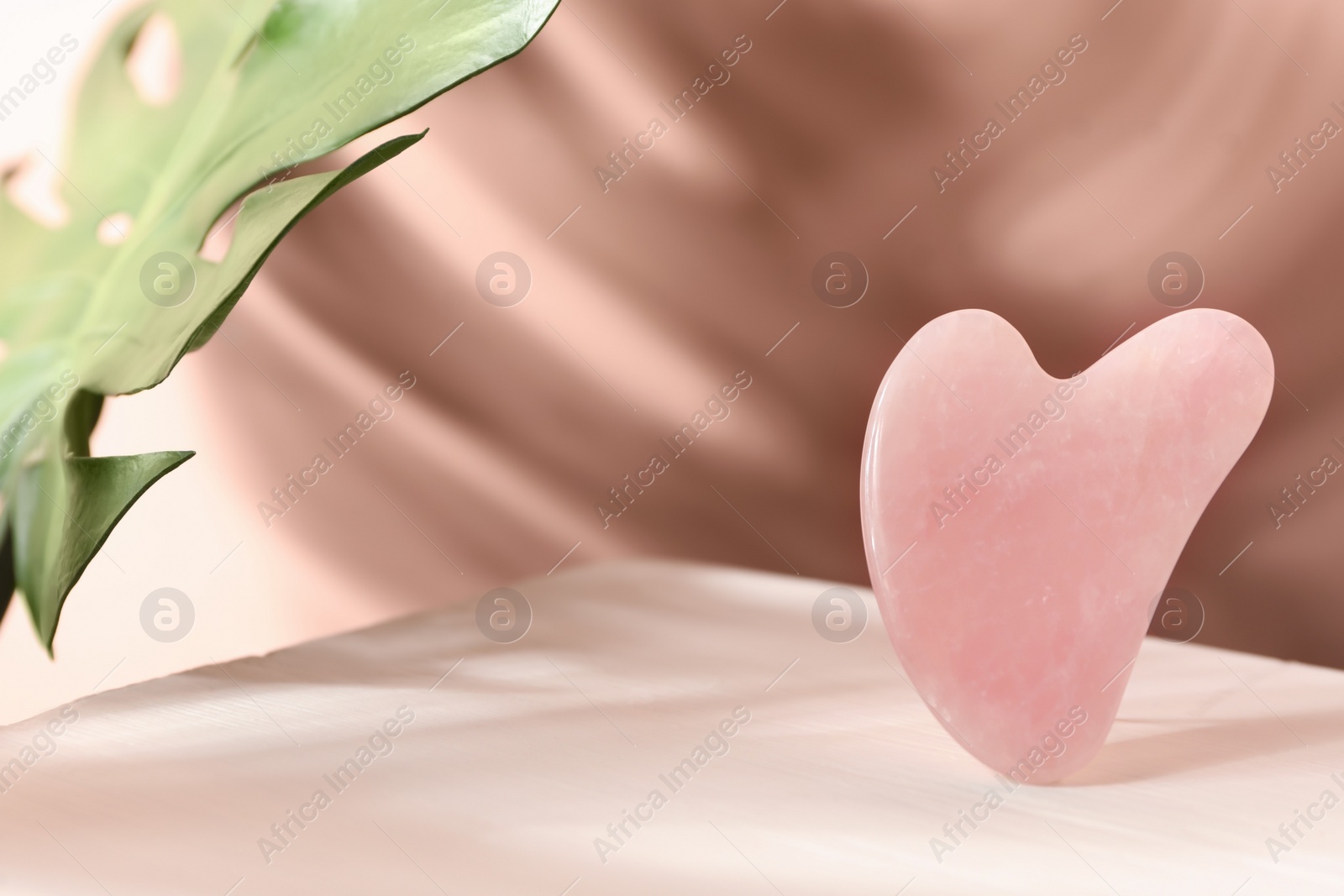 Photo of Rose quartz gua sha tool and tropical leaf on table, space for text