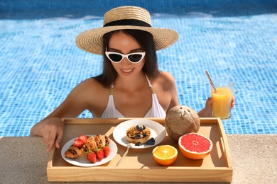 Young woman with delicious breakfast on tray in swimming pool