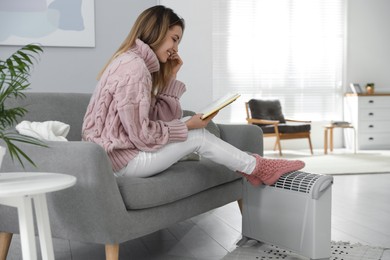 Photo of Happy young woman with book warming feet on electric heater at home