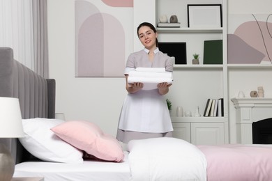 Photo of Young maid with stack of towels near bed in hotel room