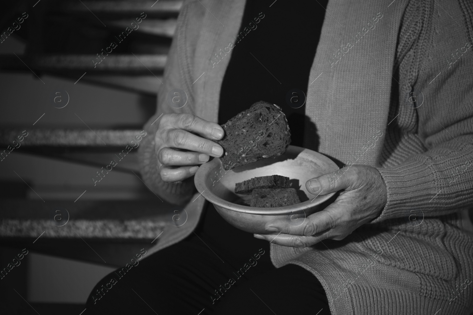 Photo of Poor senior woman with bread sitting on stairs, closeup. Black and white effect