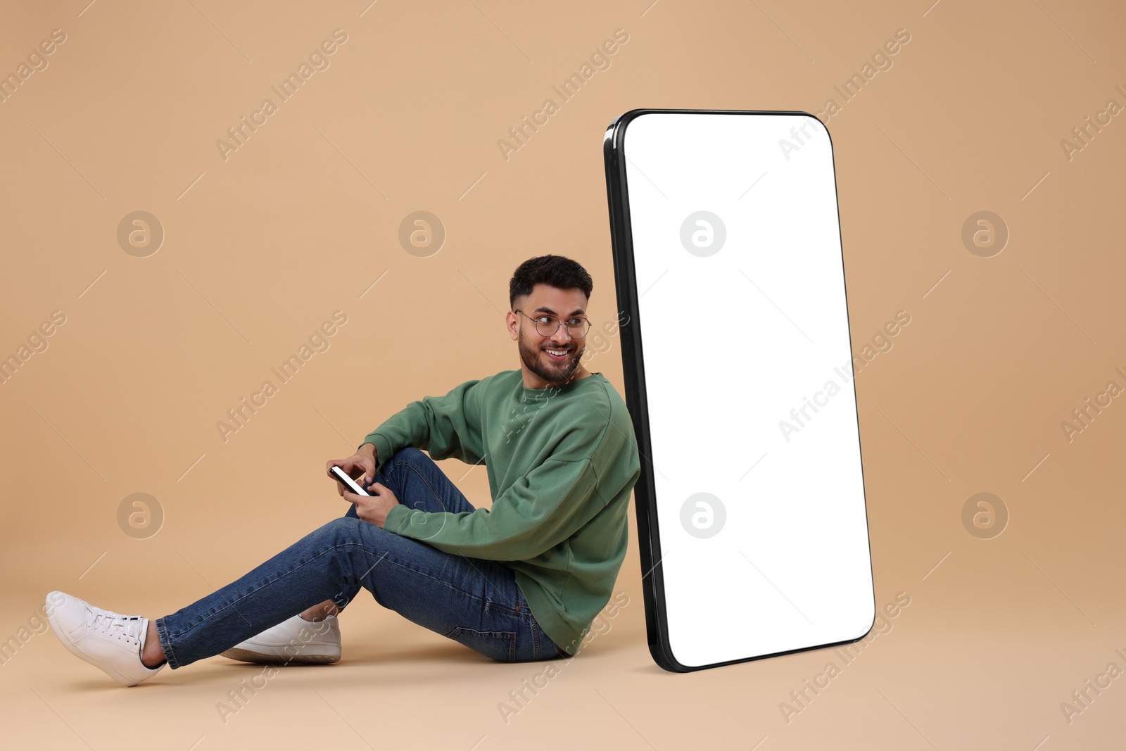 Image of Man with mobile phone sitting near huge device with empty screen on dark beige background. Mockup for design