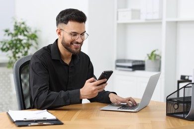 Photo of Handsome young man using smartphone while working with laptop at wooden table in office