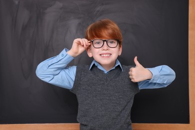 Photo of Smiling schoolboy showing thumb up near blackboard