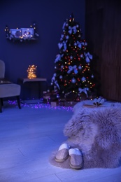 Photo of Soft slippers with fluffy blanket and Christmas tree in dark room