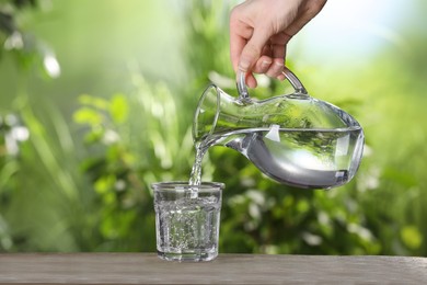 Woman pouring water from jug into glass on wooden table outdoors, closeup