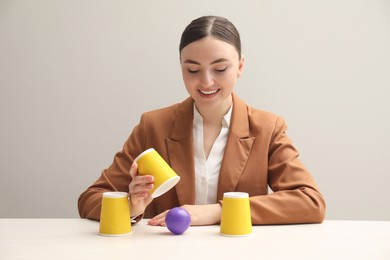 Photo of Shell game. Happy woman showing ball under cup at white table