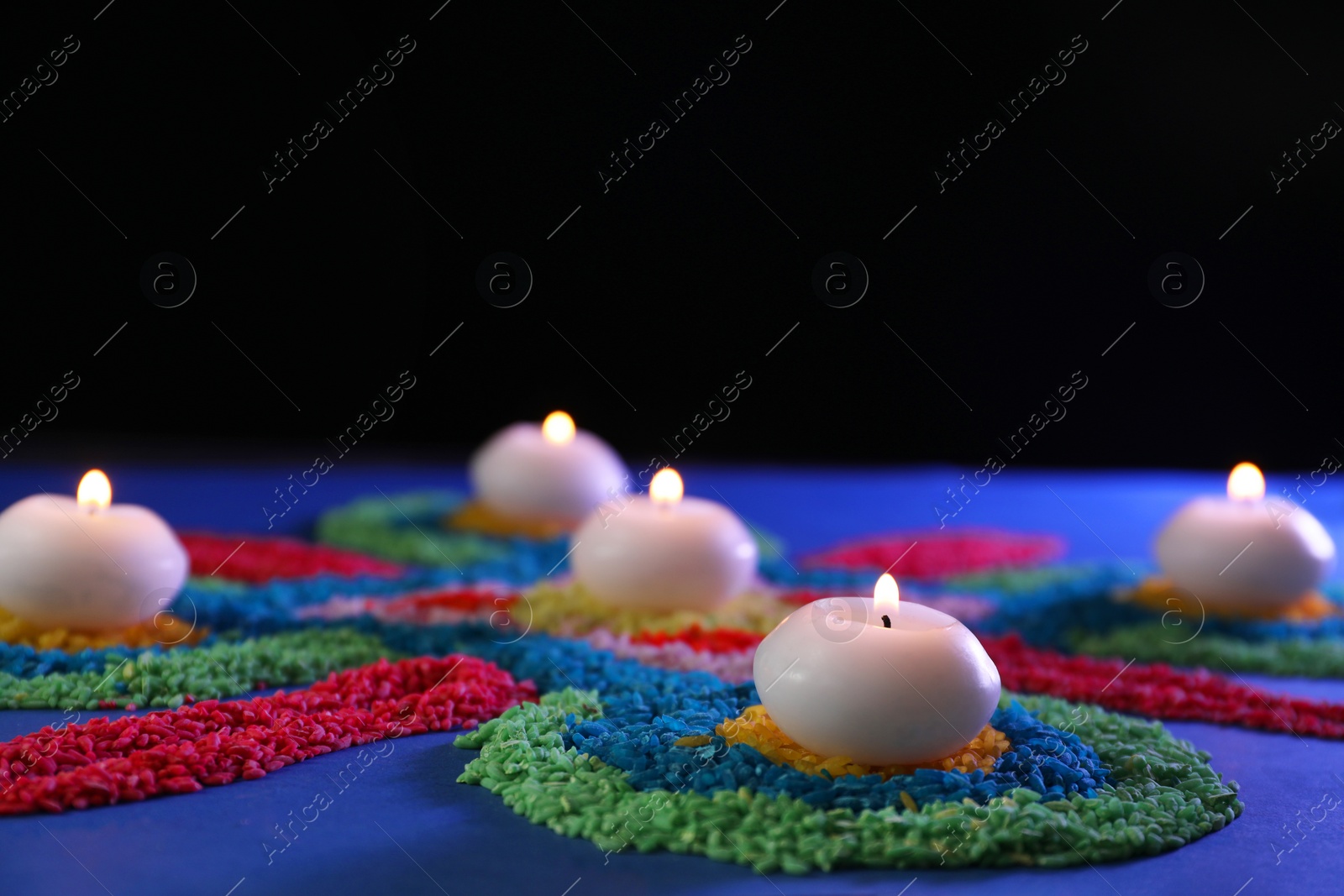 Photo of Diwali celebration. Burning candles and colorful rangoli on table against black background, closeup