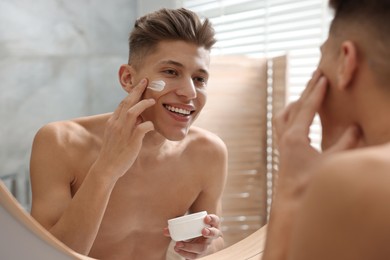 Handsome man applying moisturizing cream onto his face in bathroom