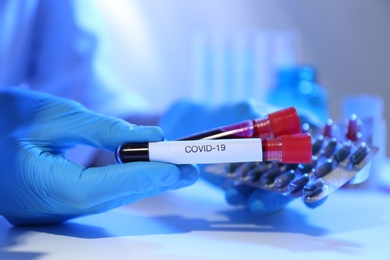 Photo of Scientist holding pills and test tubes of blood samples with label Covid-19 at table, closeup
