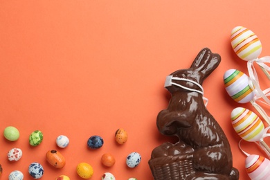 Photo of Chocolate bunny with protective mask, candies, painted eggs and space for text on coral background, flat lay. Easter holiday during COVID-19 quarantine