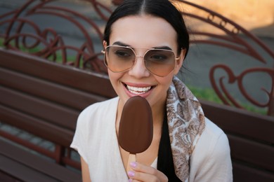 Photo of Beautiful young woman eating ice cream glazed in chocolate on bench outdoors