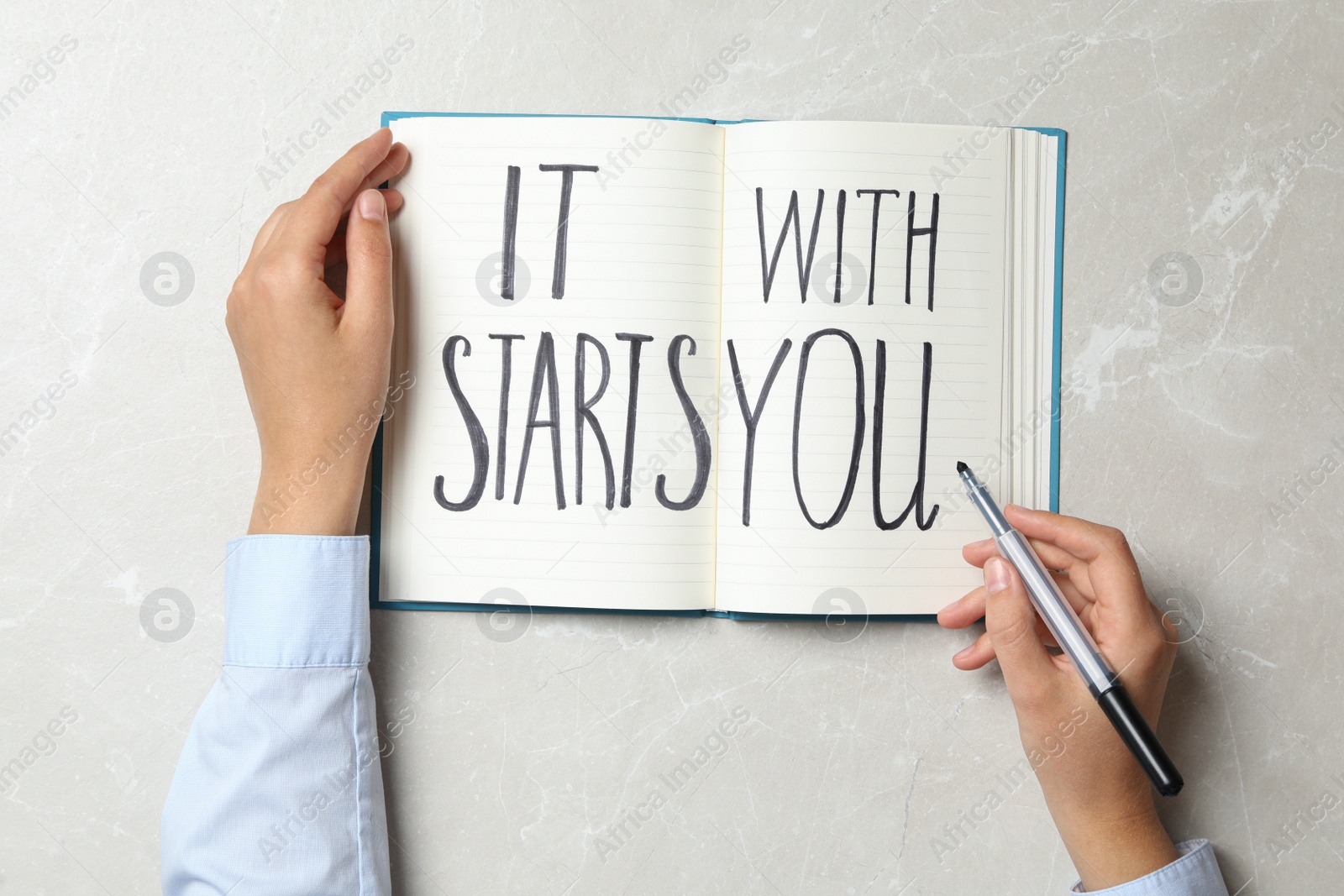 Photo of Woman writing phrase It Starts With You in notebook on grey background, top view