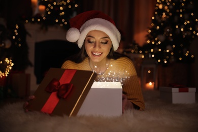 Photo of Young woman wearing Santa hat opening Christmas gift on floor at home