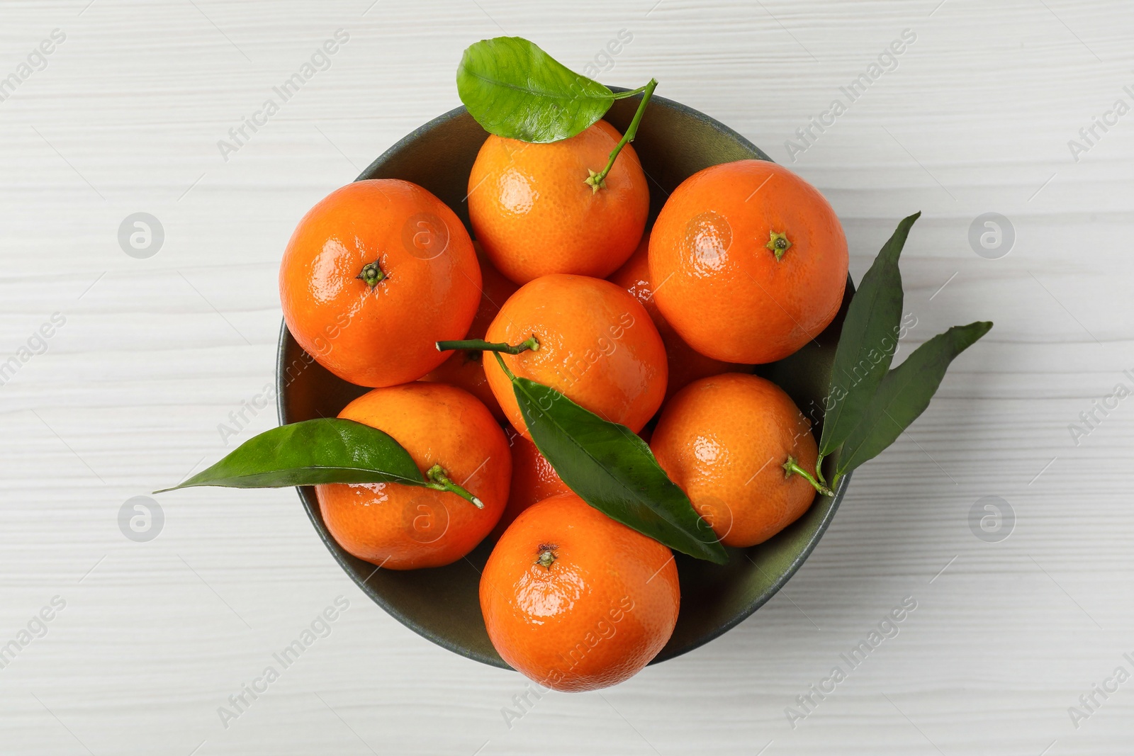 Photo of Delicious tangerines with green leaves in bowl on white wooden table, top view