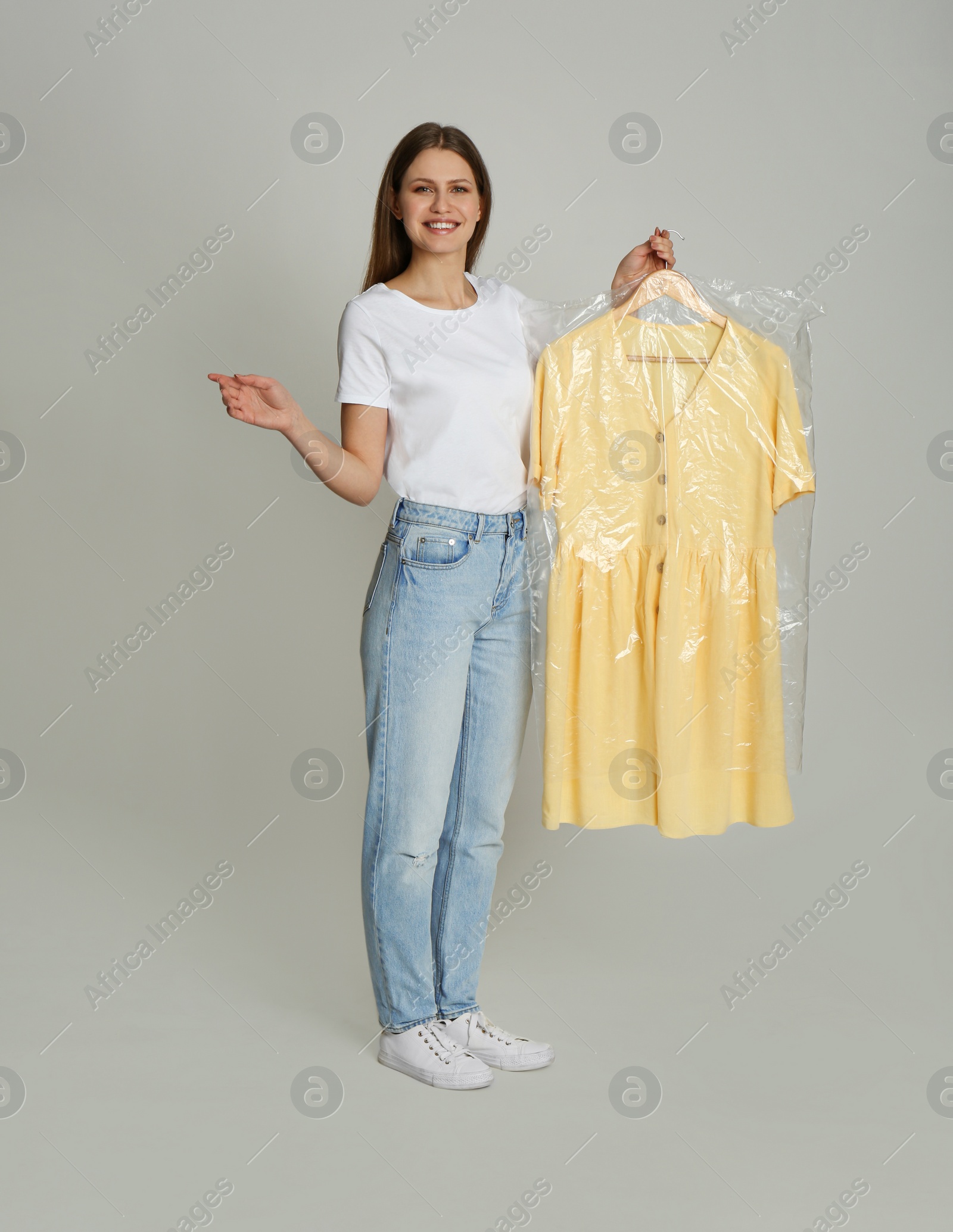 Photo of Young woman holding hanger with dress in plastic bag on light grey background. Dry-cleaning service