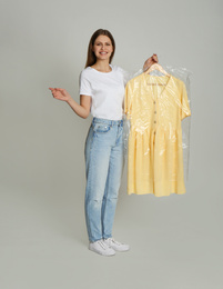 Young woman holding hanger with dress in plastic bag on light grey background. Dry-cleaning service