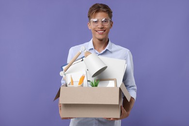 Photo of Happy unemployed young man with box of personal office belongings on purple background