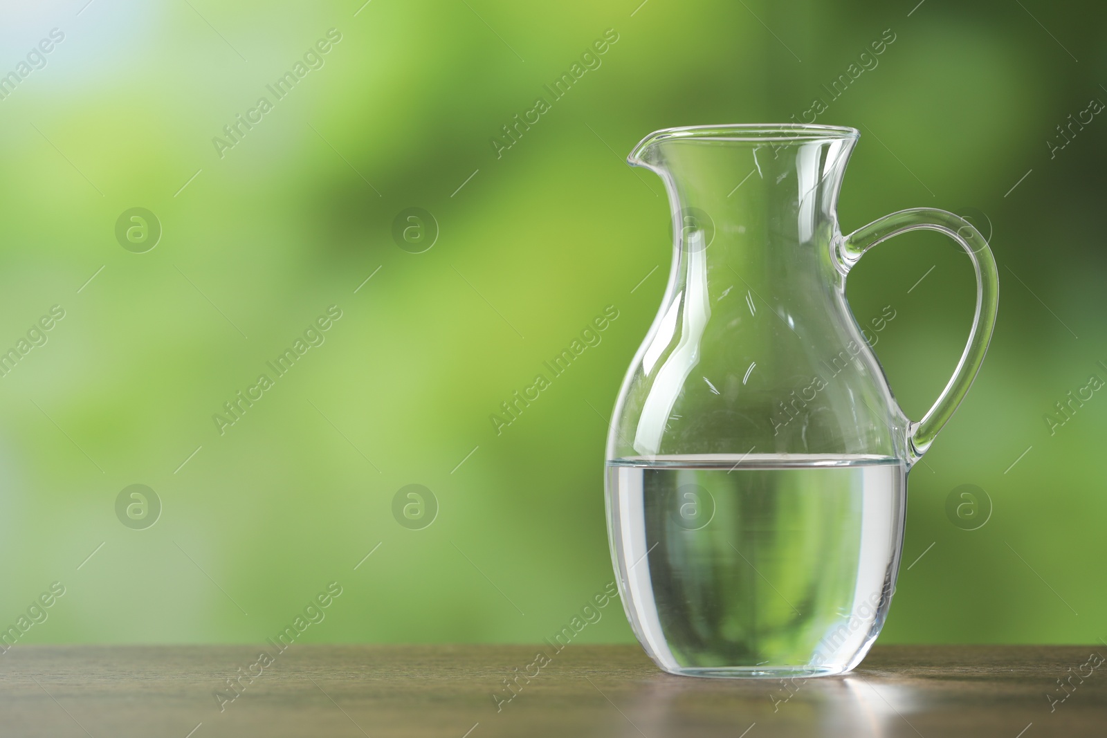 Photo of Glass jug with clear water on wooden table against blurred green background, closeup. Space for text