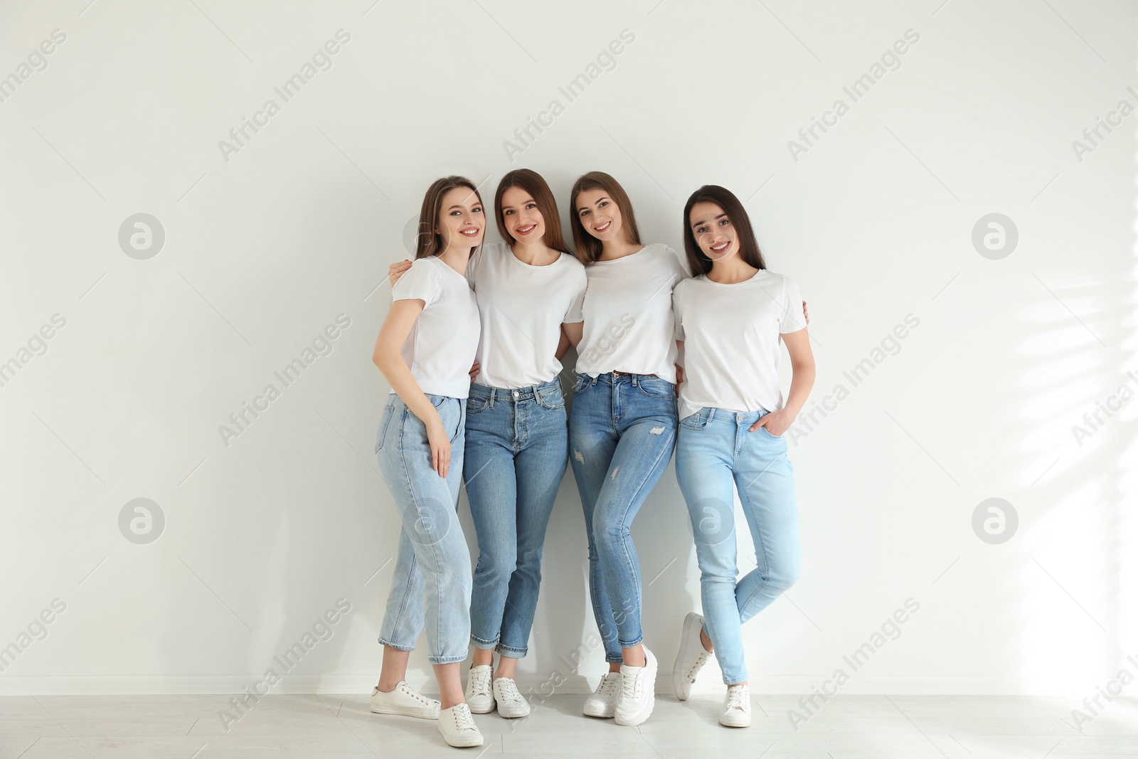 Photo of Beautiful young ladies in jeans and white t-shirts near light wall indoors. Woman's Day