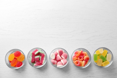 Glass bowls of different jelly candies on white wooden table, flat lay with space for text