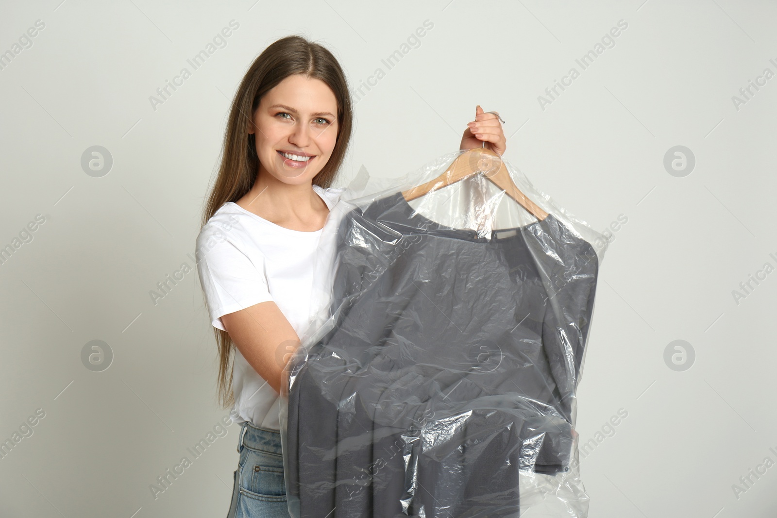 Photo of Young woman holding hanger with dress in plastic bag on light grey background. Dry-cleaning service