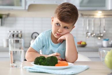 Photo of Unhappy little boy with plate of vegetables at table at table