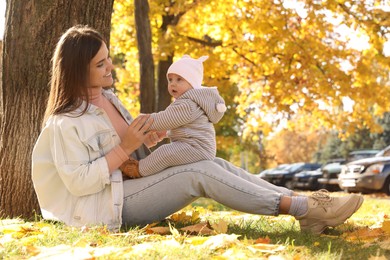 Happy mother with her baby daughter sitting near tree in park on sunny autumn day, space for text