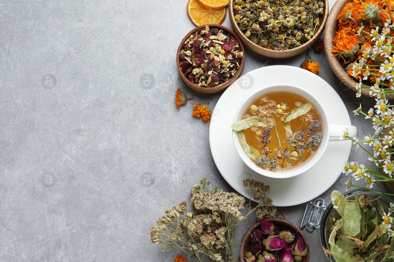 Photo of Cup of aromatic freshly brewed tea surrounded by different dry herbs on grey table, flat lay. Space for text