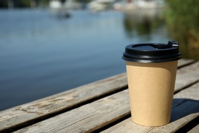 Takeaway cardboard coffee cup with plastic lid on wooden pier near river, space for text