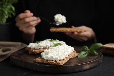 Photo of Woman spreading cottage cheese onto crispy cracker at black table, focus on board with sandwiches