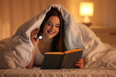 Photo of Young woman with flashlight reading book under blanket in bedroom
