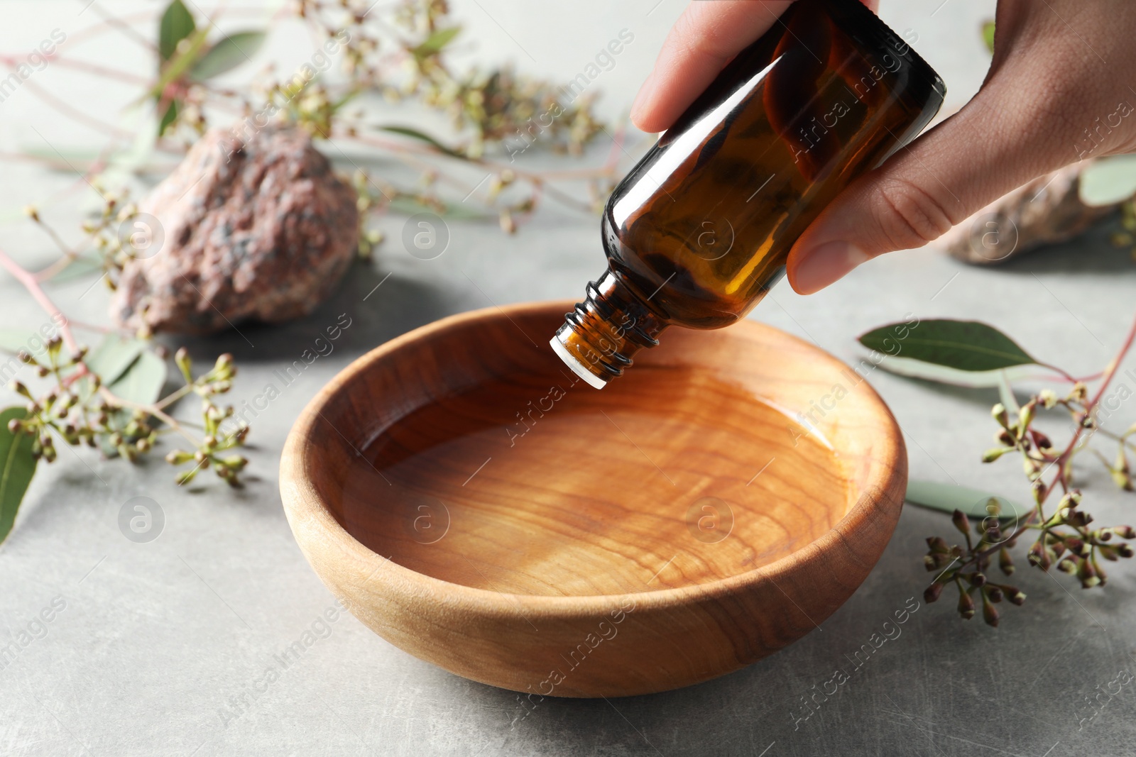 Photo of Woman dripping eucalyptus essential oil from bottle into bowl at light grey table, closeup